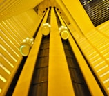 A dynamic perspective looking upward at an interior elevator system with several rounded elevator cars ascending and descending along vertical tracks. The architecture features geometric patterns and a warm yellow lighting that fills the space.