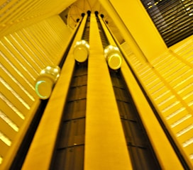A dynamic perspective looking upward at an interior elevator system with several rounded elevator cars ascending and descending along vertical tracks. The architecture features geometric patterns and a warm yellow lighting that fills the space.