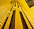 A dynamic perspective looking upward at an interior elevator system with several rounded elevator cars ascending and descending along vertical tracks. The architecture features geometric patterns and a warm yellow lighting that fills the space.