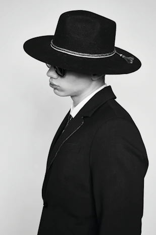 A person dressed in a dark suit and a wide-brimmed hat, wearing sunglasses. The attire is formal and the hat has a decorative band.