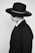 A person dressed in a dark suit and a wide-brimmed hat, wearing sunglasses. The attire is formal and the hat has a decorative band.