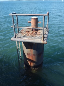 A weathered metal structure stands in the water, featuring a rusty platform with safety railing and a vertical cylindrical support. The water is a deep blue, and the surface shows gentle ripples. A metal ladder is attached to the side of the structure, descending into the water.