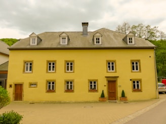 A two-story yellow building with a grey sloped roof featuring multiple dormer windows. The facade has symmetrically arranged windows, each framed in light brown with flower boxes beneath the ground floor windows. A wooden door is centrally positioned, flanked by potted plants. The building is situated on a paved area with surrounding greenery and trees visible in the background.