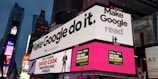 A bustling urban setting with large digital billboards displaying colorful advertisements. Prominent ads include 'Make Google do it' and 'American Idol Winner David Cook'. Bright neon colors and giant screens dominate the scene, hinting at a lively nightlife area.