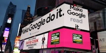 A bustling urban setting with large digital billboards displaying colorful advertisements. Prominent ads include 'Make Google do it' and 'American Idol Winner David Cook'. Bright neon colors and giant screens dominate the scene, hinting at a lively nightlife area.