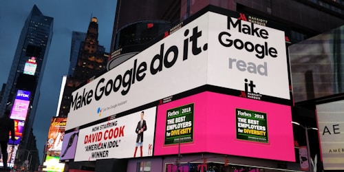 A bustling urban setting with large digital billboards displaying colorful advertisements. Prominent ads include 'Make Google do it' and 'American Idol Winner David Cook'. Bright neon colors and giant screens dominate the scene, hinting at a lively nightlife area.