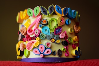 A colorful array of rolled paper art arranged on a cylindrical surface, featuring intricate designs with vibrant hues of pink, blue, yellow, green, and black. The artistic rolls create a textured and dynamic composition.