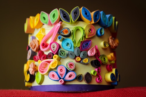 A colorful array of rolled paper art arranged on a cylindrical surface, featuring intricate designs with vibrant hues of pink, blue, yellow, green, and black. The artistic rolls create a textured and dynamic composition.
