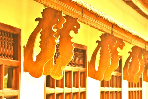 Wooden architectural elements featuring intricate scrollwork and carved designs are arranged in a pattern along the outer façade of a building. The warm tones of the wood contrast with the lighter walls, enhancing the decorative detail. The carvings suggest a traditional style, possibly influenced by cultural motifs.