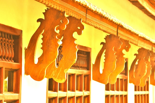 Wooden architectural elements featuring intricate scrollwork and carved designs are arranged in a pattern along the outer façade of a building. The warm tones of the wood contrast with the lighter walls, enhancing the decorative detail. The carvings suggest a traditional style, possibly influenced by cultural motifs.