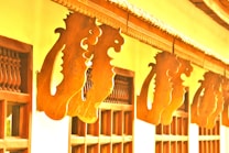 Wooden architectural elements featuring intricate scrollwork and carved designs are arranged in a pattern along the outer fa&ccedil;ade of a building. The warm tones of the wood contrast with the lighter walls, enhancing the decorative detail. The carvings suggest a traditional style, possibly influenced by cultural motifs.