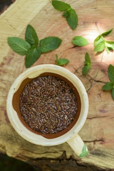 A cream-colored ceramic cup filled with brown flaxseeds rests upon a wooden surface. Surrounding the cup are fresh green mint leaves scattered across the wood, which displays a natural, rustic texture.