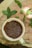 A cream-colored ceramic cup filled with brown flaxseeds rests upon a wooden surface. Surrounding the cup are fresh green mint leaves scattered across the wood, which displays a natural, rustic texture.