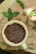 A cream-colored ceramic cup filled with brown flaxseeds rests upon a wooden surface. Surrounding the cup are fresh green mint leaves scattered across the wood, which displays a natural, rustic texture.