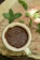 A cream-colored ceramic cup filled with brown flaxseeds rests upon a wooden surface. Surrounding the cup are fresh green mint leaves scattered across the wood, which displays a natural, rustic texture.