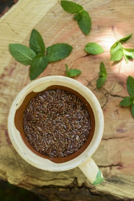 A cream-colored ceramic cup filled with brown flaxseeds rests upon a wooden surface. Surrounding the cup are fresh green mint leaves scattered across the wood, which displays a natural, rustic texture.