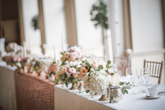 A beautifully arranged wedding table setting featuring elegant floral centerpieces with roses and hydrangeas in soft pastel colors. The table is adorned with glassware, a teacup, and decorative candle holders, creating an elegant and romantic atmosphere. The blurred background suggests a spacious, well-lit room with large windows.
