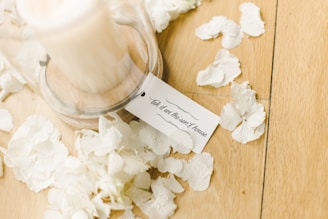 A candle under a glass cloche sits atop a wooden surface, surrounded by white flower petals. A small card with elegant script is placed near the base of the cloche.