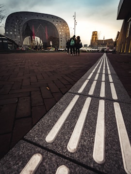 A modern architectural building with a large, reflective arched window stands prominently in the background. The foreground features a textured pathway with tactile strips. Several people are walking in the distance, and the scene is set during an overcast day with a slight glow from the setting or rising sun.