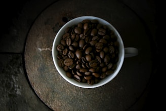 A white cup filled to the brim with dark roasted coffee beans sits on a rustic, circular stone table. The dim lighting creates a moody and cozy atmosphere, highlighting the rich texture and shine of the beans.