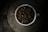 A white cup filled to the brim with dark roasted coffee beans sits on a rustic, circular stone table. The dim lighting creates a moody and cozy atmosphere, highlighting the rich texture and shine of the beans.