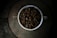 A white cup filled to the brim with dark roasted coffee beans sits on a rustic, circular stone table. The dim lighting creates a moody and cozy atmosphere, highlighting the rich texture and shine of the beans.
