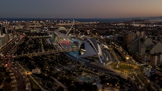 An aerial view of a modern architectural complex at dusk, with sleek, futuristic buildings and a well-lit, expansive urban landscape. The sky is dimly lit, and city lights illuminate the structures and nearby roads. Surrounding the architecture, there are landscaped areas with greenery and water features.