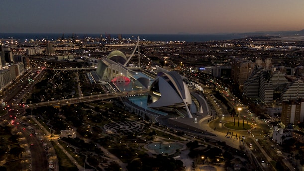 An aerial view of a modern architectural complex at dusk, with sleek, futuristic buildings and a well-lit, expansive urban landscape. The sky is dimly lit, and city lights illuminate the structures and nearby roads. Surrounding the architecture, there are landscaped areas with greenery and water features.