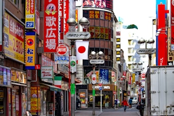 A bustling urban street scene filled with vibrant storefront signage, including Japanese text and logos, large colorful billboards, and promotional posters. Numerous signs advertise restaurants, shops, and businesses, with pedestrians walking along the sidewalk. A Japanese flag is prominently displayed in the middle of the scene.
