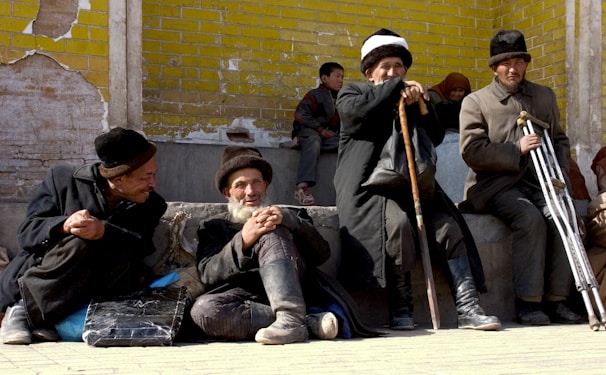 A group of older men wearing traditional hats and clothing sit on the ground in front of a yellow brick wall. They are engaged in conversation, holding walking sticks. In the background, children sit lined against the wall, observing quietly.