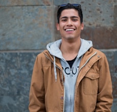 man in brown coat standing near concrete wall smiles