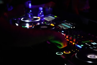 A DJ is mixing music at a dimly lit nightclub, surrounded by a variety of glowing and illuminated buttons and dials on a mixing console. The vibrant lights from the equipment contrast with the dark environment, and there is a sense of motion as the DJ adjusts the controls.