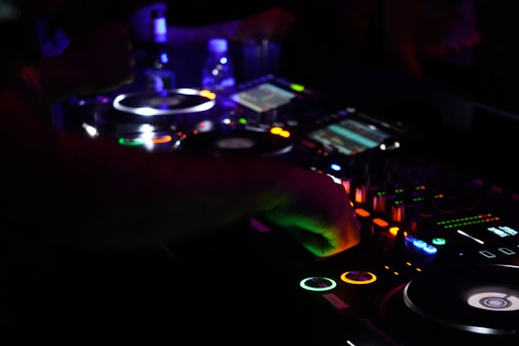 A DJ is mixing music at a dimly lit nightclub, surrounded by a variety of glowing and illuminated buttons and dials on a mixing console. The vibrant lights from the equipment contrast with the dark environment, and there is a sense of motion as the DJ adjusts the controls.