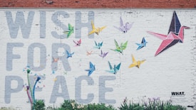 A mural on a white brick wall features a large phrase 'WISH FOR PEACE' in light gray letters. Colorful origami cranes of different sizes and colors, such as blue, green, yellow, and pink, are depicted flying around. There are also small colorful plants at the bottom of the wall.