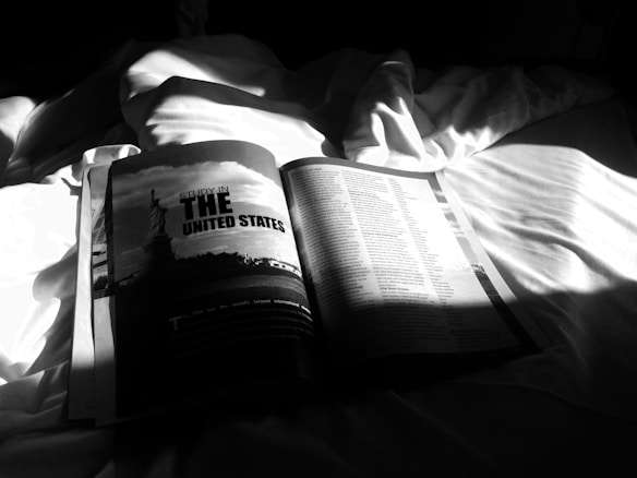 An open magazine lies on a rumpled bed with bright sunlight casting shadows across its pages. The visible page headline reads 'Study in the United States' alongside an image of the Statue of Liberty. Additional text is printed on the right-hand side with a visually dark and dramatic light contrast.