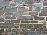 A wall constructed from various sizes and shapes of stacked stones, with visible mortar between them. The stones have a rough, textured surface and a mix of dark and light gray tones.