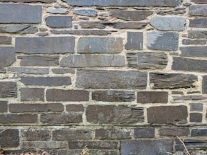 A wall constructed from various sizes and shapes of stacked stones, with visible mortar between them. The stones have a rough, textured surface and a mix of dark and light gray tones.