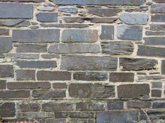 A wall constructed from various sizes and shapes of stacked stones, with visible mortar between them. The stones have a rough, textured surface and a mix of dark and light gray tones.