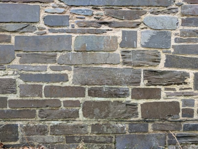 A wall constructed from various sizes and shapes of stacked stones, with visible mortar between them. The stones have a rough, textured surface and a mix of dark and light gray tones.