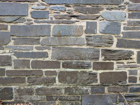A wall constructed from various sizes and shapes of stacked stones, with visible mortar between them. The stones have a rough, textured surface and a mix of dark and light gray tones.