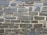 A wall constructed from various sizes and shapes of stacked stones, with visible mortar between them. The stones have a rough, textured surface and a mix of dark and light gray tones.