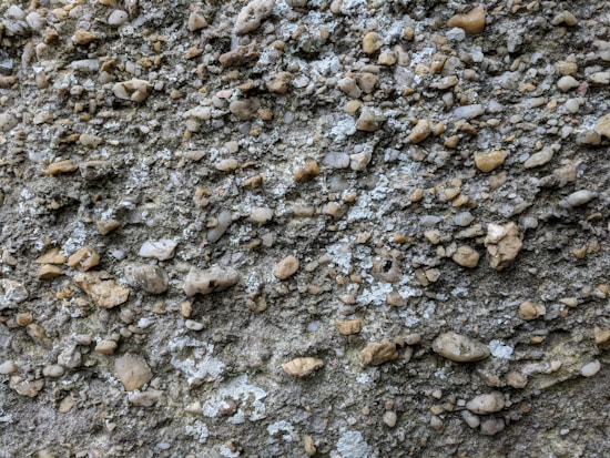 A close-up view of a textured surface composed of numerous small, irregularly shaped stones embedded in a gritty matrix. The stones vary in size and color, creating a natural, rugged appearance. The surface shows a mixture of gray, tan, and some whitish elements.