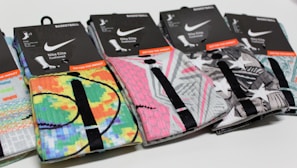 A row of colorful Nike Elite basketball socks with various graphic designs and packaging labels. The designs include bright patterns and abstract shapes, with each pair featuring a black line detail. The packaging is predominantly black with white and orange text.
