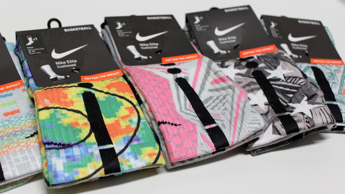 A row of colorful Nike Elite basketball socks with various graphic designs and packaging labels. The designs include bright patterns and abstract shapes, with each pair featuring a black line detail. The packaging is predominantly black with white and orange text.