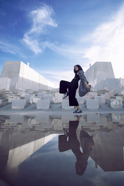 A person is walking confidently on a concrete surface with a dynamic pose, casting a vivid reflection on a mirror-like puddle below. The background features stark modern architecture with white and gray geometric shapes under a vibrant blue sky with scattered clouds.