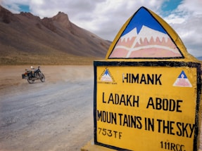 A road sign with the text 'Ladakh Abode Mountains in the Sky' stands prominently in a barren, mountainous landscape. A motorcycle is parked on a dusty road, and rugged hills rise in the background under a partly cloudy sky.