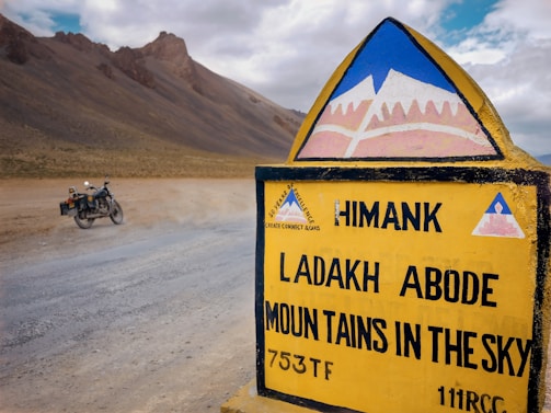 A road sign with the text 'Ladakh Abode Mountains in the Sky' stands prominently in a barren, mountainous landscape. A motorcycle is parked on a dusty road, and rugged hills rise in the background under a partly cloudy sky.