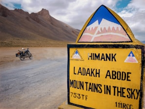 A road sign with the text 'Ladakh Abode Mountains in the Sky' stands prominently in a barren, mountainous landscape. A motorcycle is parked on a dusty road, and rugged hills rise in the background under a partly cloudy sky.