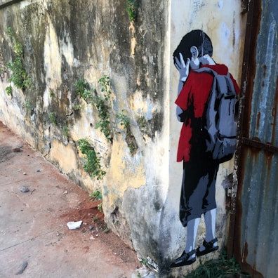 A piece of street art portraying a child leaning against an aged, weathered wall with patches of green foliage. The child is depicted in a stylized manner, wearing a red shirt and a backpack, with hands covering their face as if in sadness or distress. The wall and ground show signs of decay and neglect.
