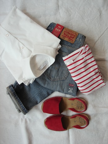A set of clothing items are neatly arranged on a white background, including a folded white dress shirt, a pair of blue denim jeans with a visible Levi's label, a red and white striped shirt, and a pair of red open-toe sandals.